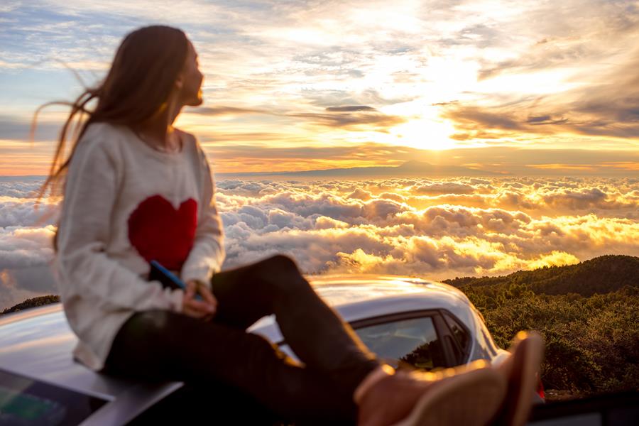 A person with long hair, a white jumper with a red heart on it and black pants and brown shoes. They are sitting on the roof of a car looking out over clouds during a sunset. Its a beautiful view.