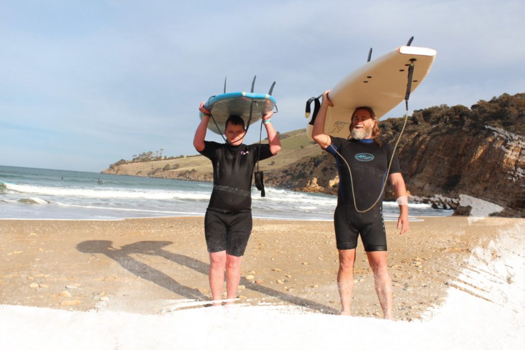 2 people on the beach walking out of the surf wearing wetsuits and with surfboards rested on their heads.