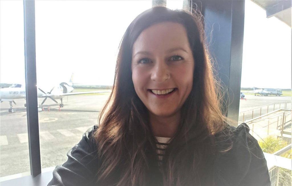 Once a month Jade Rhodes flies to King Island to provide outreach support to local families. Jade is sitting at the airport and you can see a small aeroplane in the background. She is smiling.