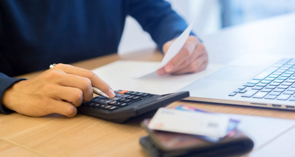 person sitting at laptop with a calculator, paper in hand and wallet sitting on table with credit cards out