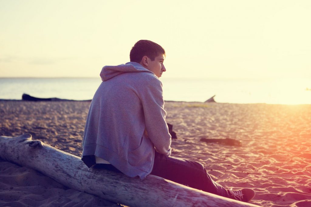 A male youth staying in crisis accommodation contemplating homelessness while sitting on the beach.