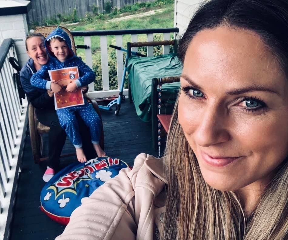 A smiling person taking a selfie with a smiling mum and child sitting in the background.