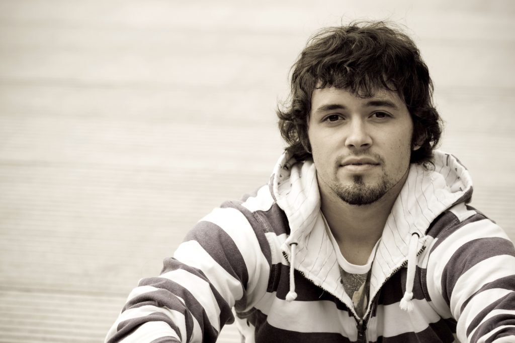 Young person with a goaty, dark hair wearing a stripy hooded jumper. Looks serious. Appears to be sitting on a roof.