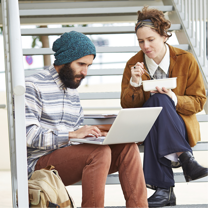 A photo of two people sittingon some stairs. One person is typing on a laptop, the other is eating their lunch and watching them.