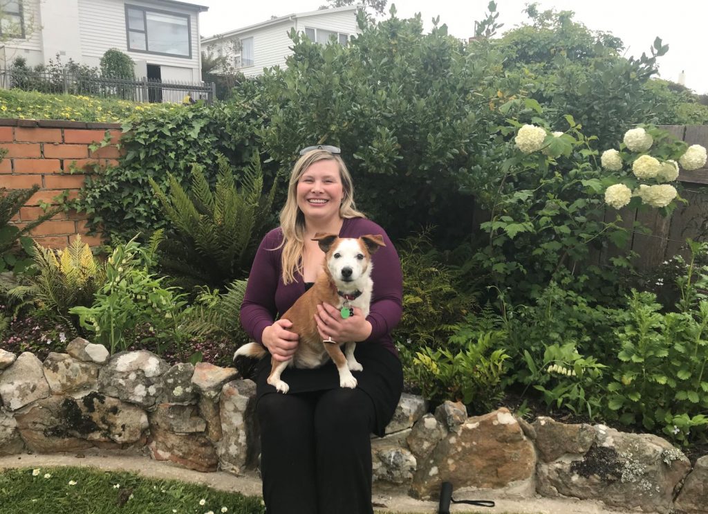 Kerrie is sitting outside in her garden with Scooter on her lap. Scooter is a gorgeous small brown and white dog with floppy ears. Kerrie has a big happy smile on her face.