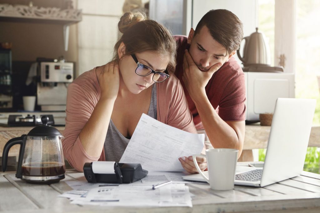 A couple at the kitchen bench standing over a bunch of papers, a laptop and a calculator looking concerned.