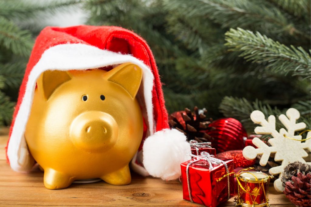 Piggybank with a santa hat on sitting in front of a christmas tree.