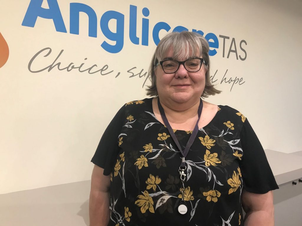 Anglicare Tasmania Financial Counsellor Fiona Moore standing and smiling. She is wearing a black shirt with flowers and black glasses. Fiona is standing in front of a wall that has the Anglicare Tasmania logo on it.