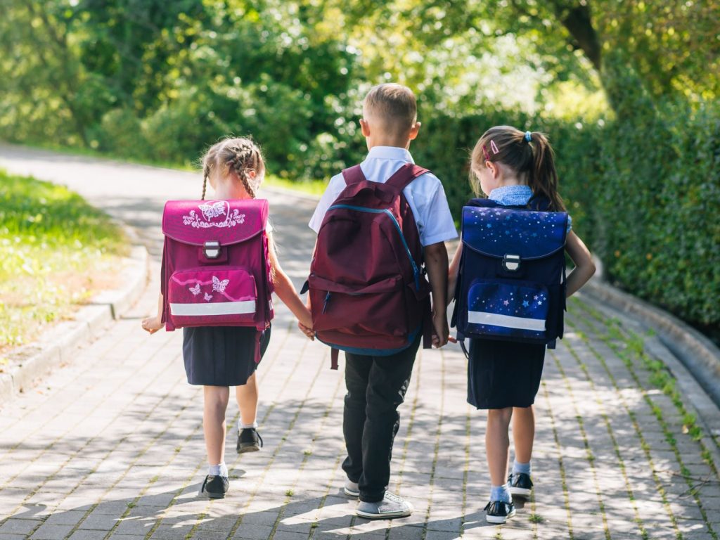 Children walking hand in hand in a school uniform with a school bag on their back.