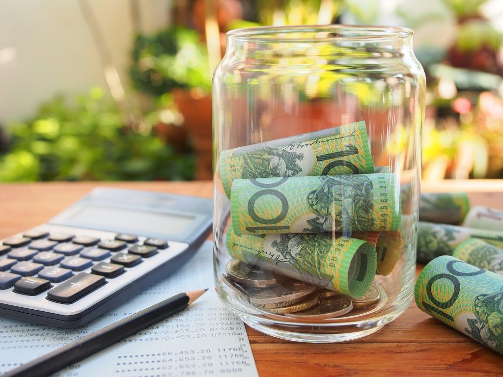 A jar with 100 dollar notes rolled up inside. Sits beside a caculator, a pencil and a piece of paper with numbers on it.