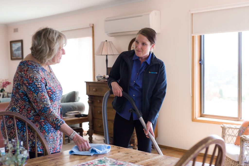 photo of an Anglicare home care support worker doing vacumning while a client is polishing their wooden dining table. They are smiling and talking as they clean.