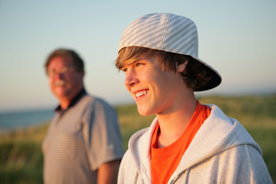 Young person with a cap on backwards, smiling, standing sidewards to the camera. I older person is standing in the background.