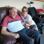 Garry sitting in his armchair and smiling widely. His support worker Donna is kneeling beside him.