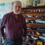 Kees is standing in front of a bookshelf full of model cars of many different types. There is also a nearby glass case full of more model cars.