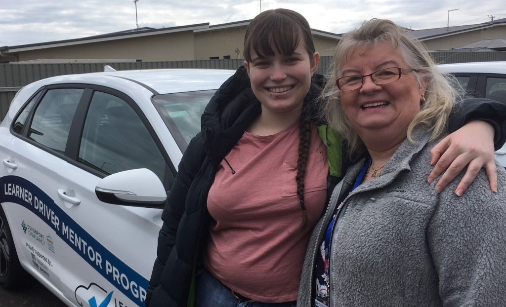 2 people, one looking younger than the other, smiling, leaning against a car as if they have been driving together.