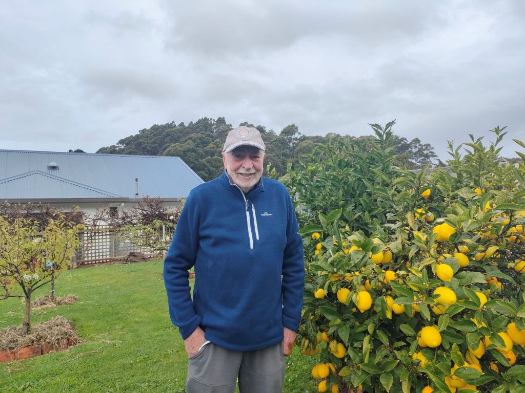 Photo of man standing in the garden beside a tree with lots of ripe lemons. He is smiling, wearing a baseball cap and has his hands in his pockets.