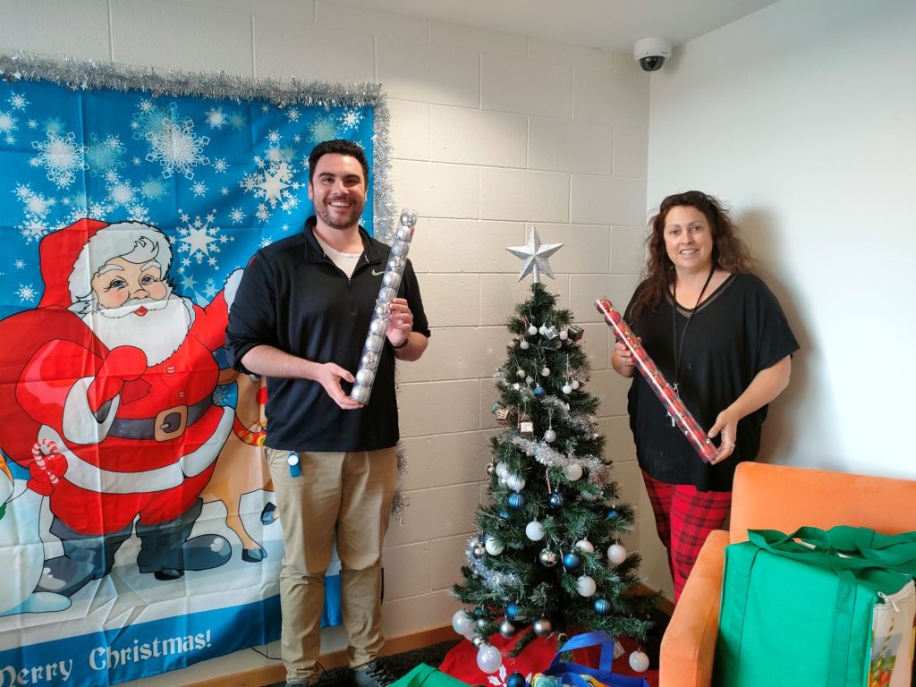 Eveline House staff members Christian and Anne - Maree are standing beside a christmas tree and a large christmas banner is on the wall with Santa Claus on it. Underneath the tree are 10 or more cooler bags.