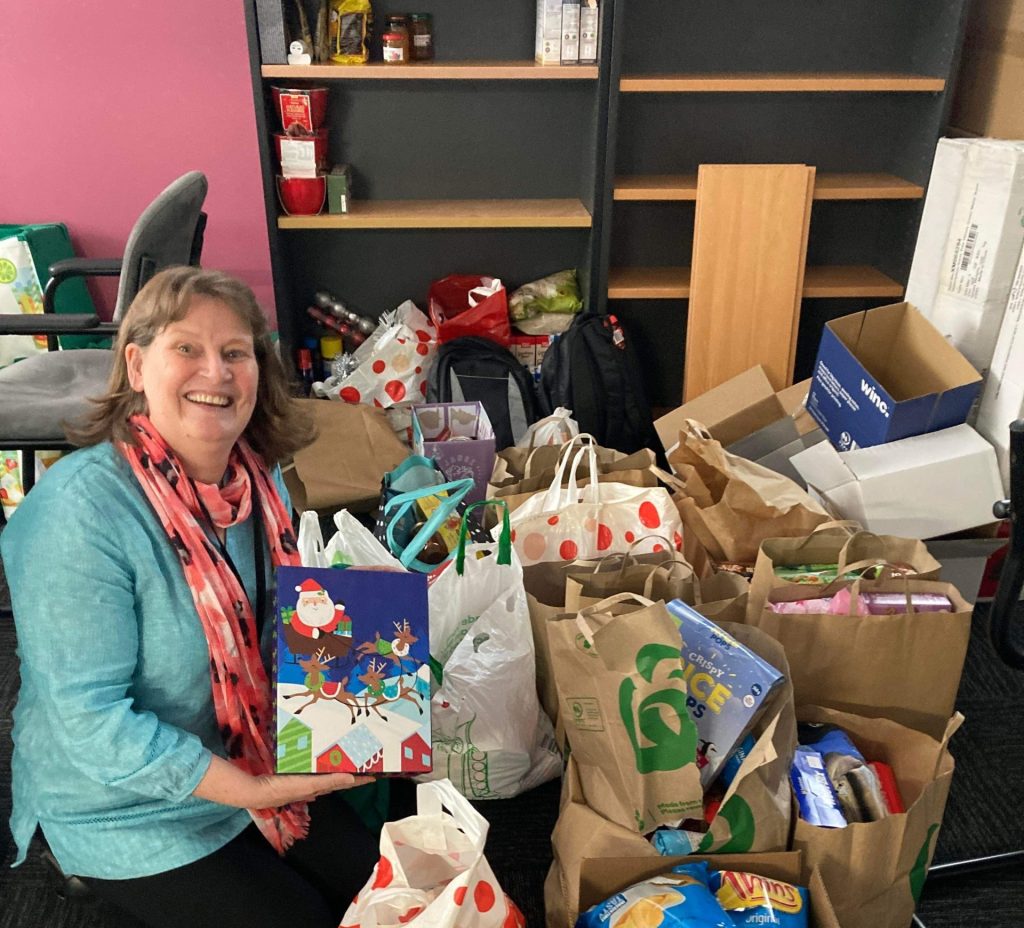 A photo of Margaret Savage amongst all the donated items for the Christmas Appeal. She looks happyily overwhelmed.