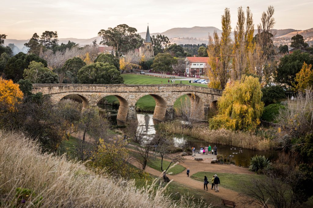 View of RIchmond Bridge, RIchmond, Southern Tasmania. Heritage bridge made from sandstone. Water running underneath. People walking along pathway beside the river and standing by the river feeding ducks. Sandstone Church in the distance. Autumn leaves on the trees.