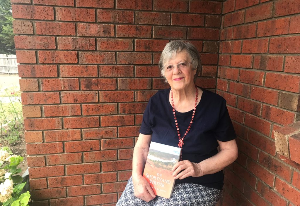 A seated, older lady displays the first book she wrote on her lap.