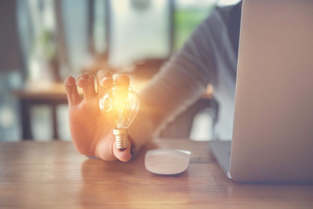 Woman sitting in front of a laptop holding a lit up lightbulb in her hand.