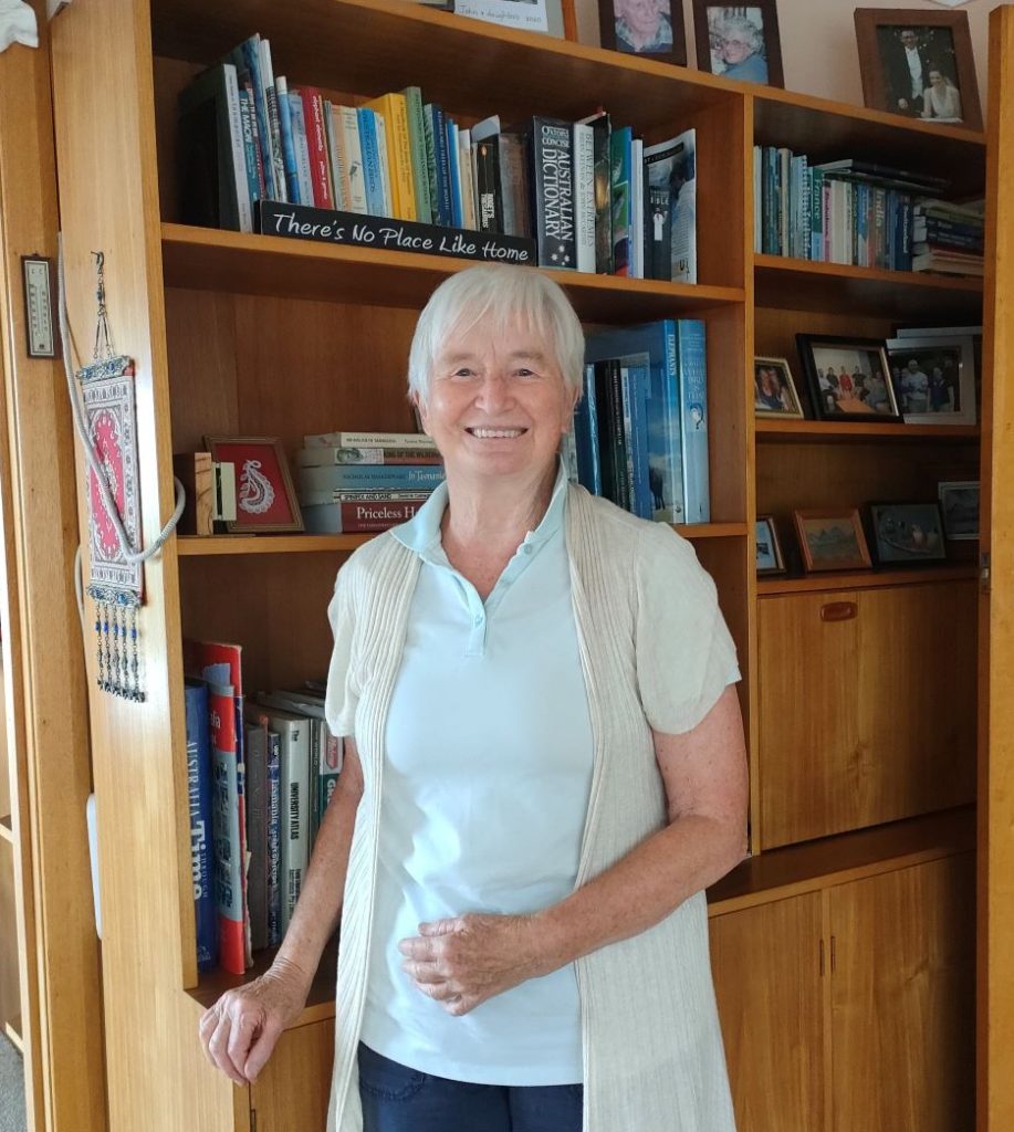 Photo of Anne Marie an acquired injury support service client. She is standing beside a bookshelf, smiling.
