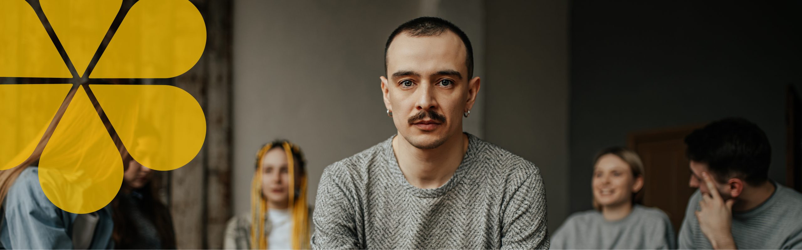 person with a moustache looking seriously at the camera and looking worried like they need some help. People sitting in the background.