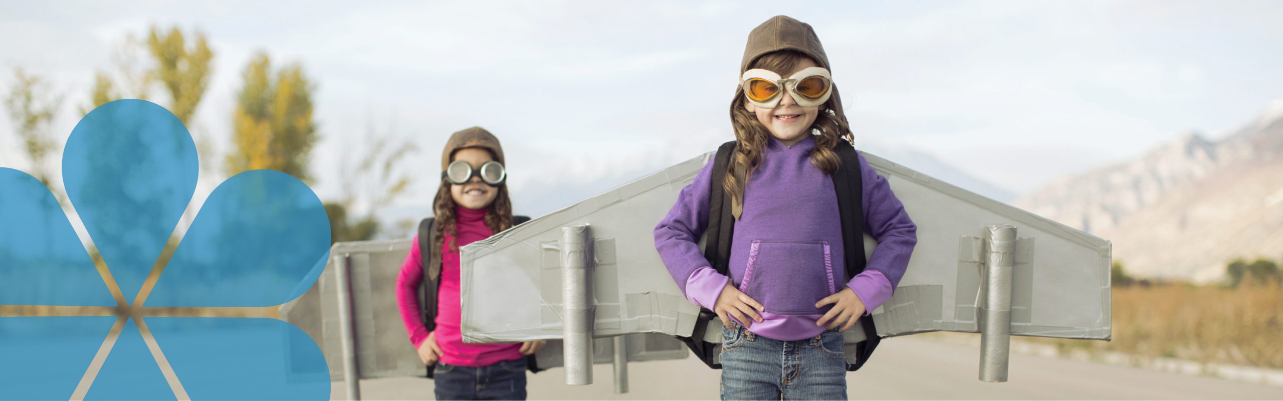 A picture of 2 young girls wearing old fashion flying hats and goggles. They have home made carboard aeroplane wings on. They look happy, smiling and confident like they are going to take on the world.