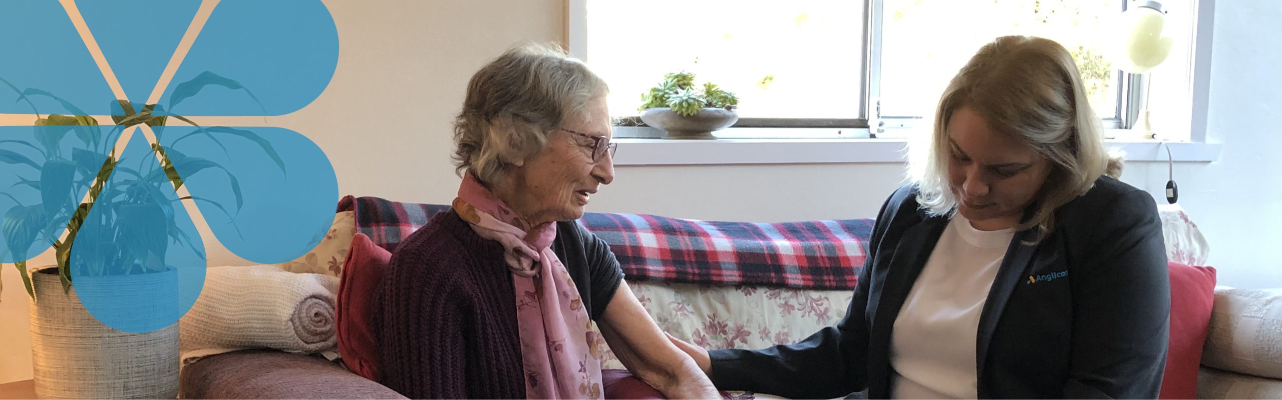Image of an Anglicare Home Care Team Member with a client sitting on the couch. Appears to be taking the clients blood pressure.