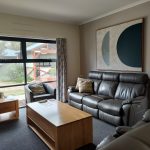 A shared lounge area at Rocherlea with leather couches, large coffee table, side table, lap, by a window and artwork on the wall.