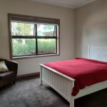 A unit bedroom at Rocherlea. It is large and carpeted, with a large window with a view of the garden. There is a timber queen sized bed, painted white. A single arm chair with a pillow.