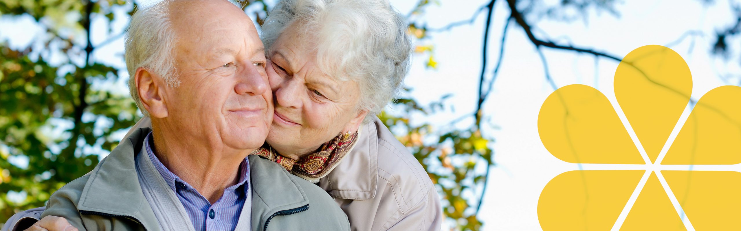 An older couple. One of them hugging the other from behind with their cheek resting on the other persons cheek. They are smiling.