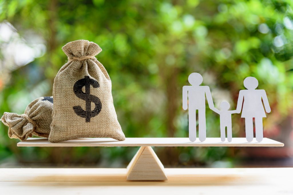 image of a balancing scale with a bag full of money on one side and a paper cutout of a family on the other.