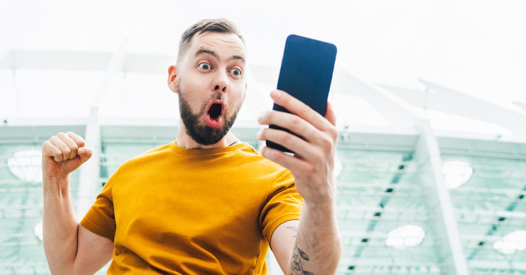 A male excitedly reacting to a wager placed on a mobile sports betting app. The male is standing outside a sports stadium.