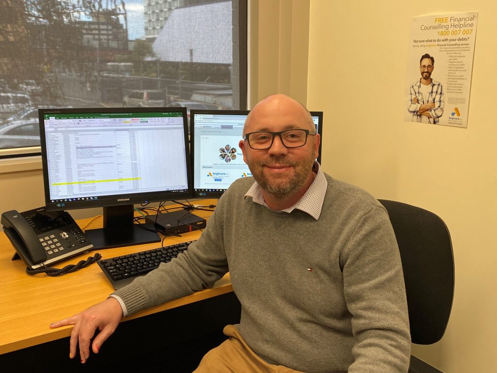 Mat O'Brien, Financial Resillience Wellbeing Program Manager sitting at his desk turned away from his computer looking at the camera similing.