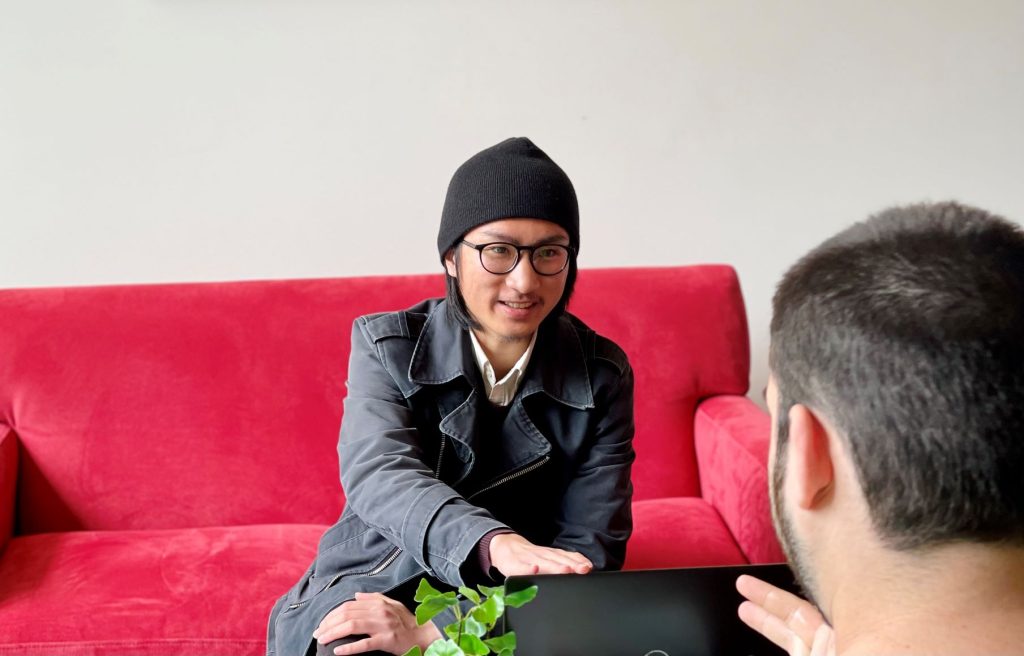 Photo of student sitting on a red couch with their laptop talking to someone sitting opposite them.