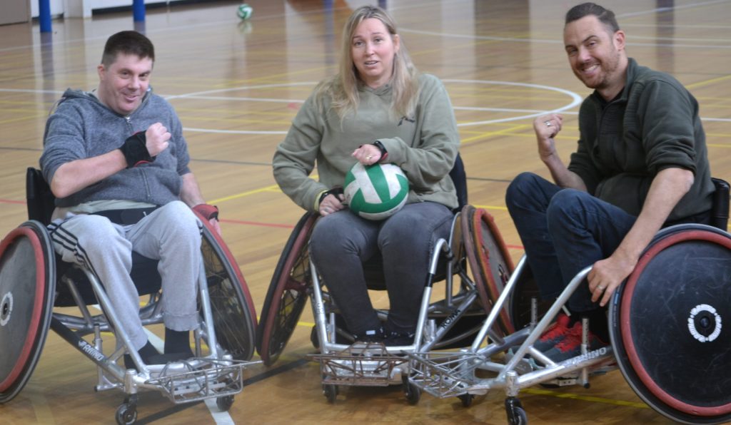 A photo of Alysse in her wheelchair playing rugby. All three people look happy and ready to play.