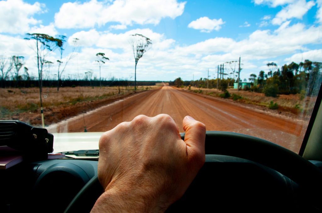 Looking out of the drivers seat of a care as it drives on an outback dirt road. All you can seek is the hand of the person driving.