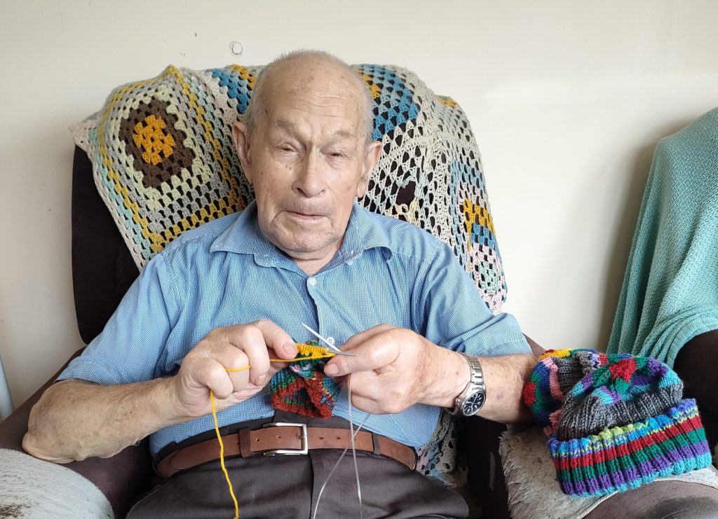 Anglicare Aged Home Care Client Keven sitting in his recliner knitting. There is a crochet rug on the seat behind him and a completed beanie on the arm of the chiar.