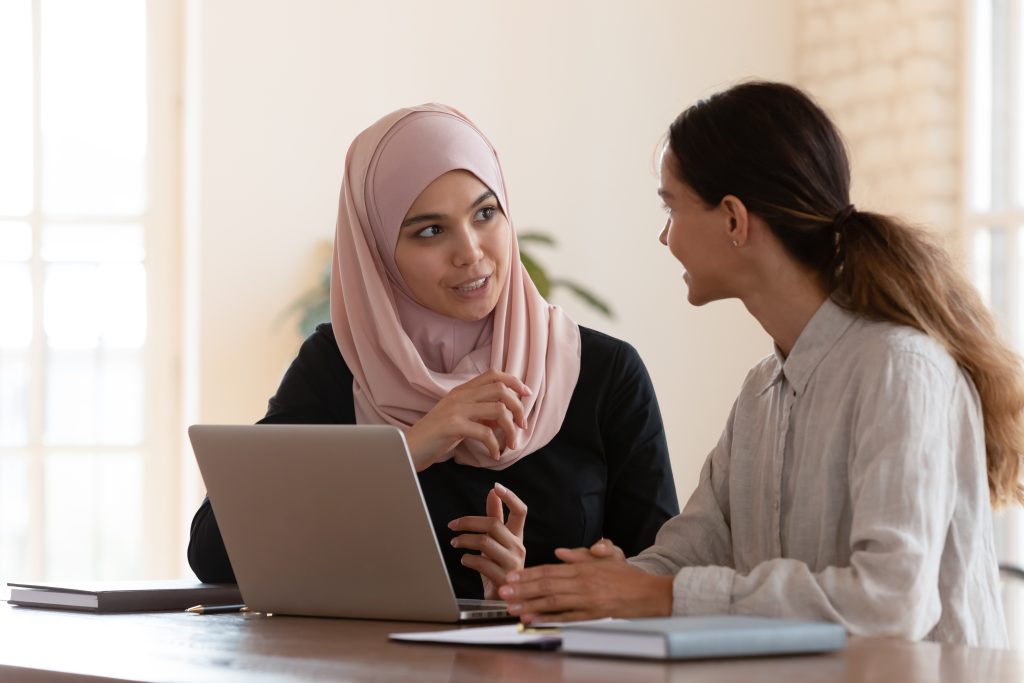 two ladies talking at a desk with a laptop in front of them.