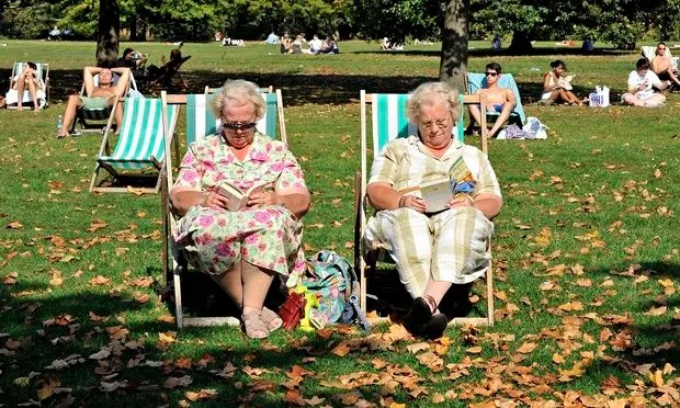 Two lovely older ladies sitting on deck chairs in the park ready books. They are wearing floral dresses and sandles. Lots of other people in the park also enjoying the sun. There are autmn leaves on the ground.