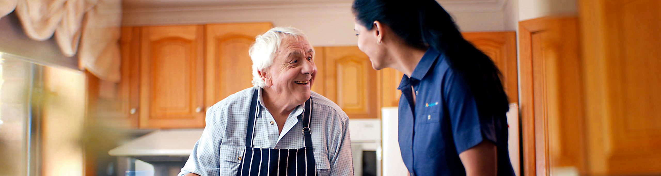 Older man in his kitchen with a support worker wearing an Anglicare Tasmania Home Care uniform. They are making a cake. They are talking and smiling.