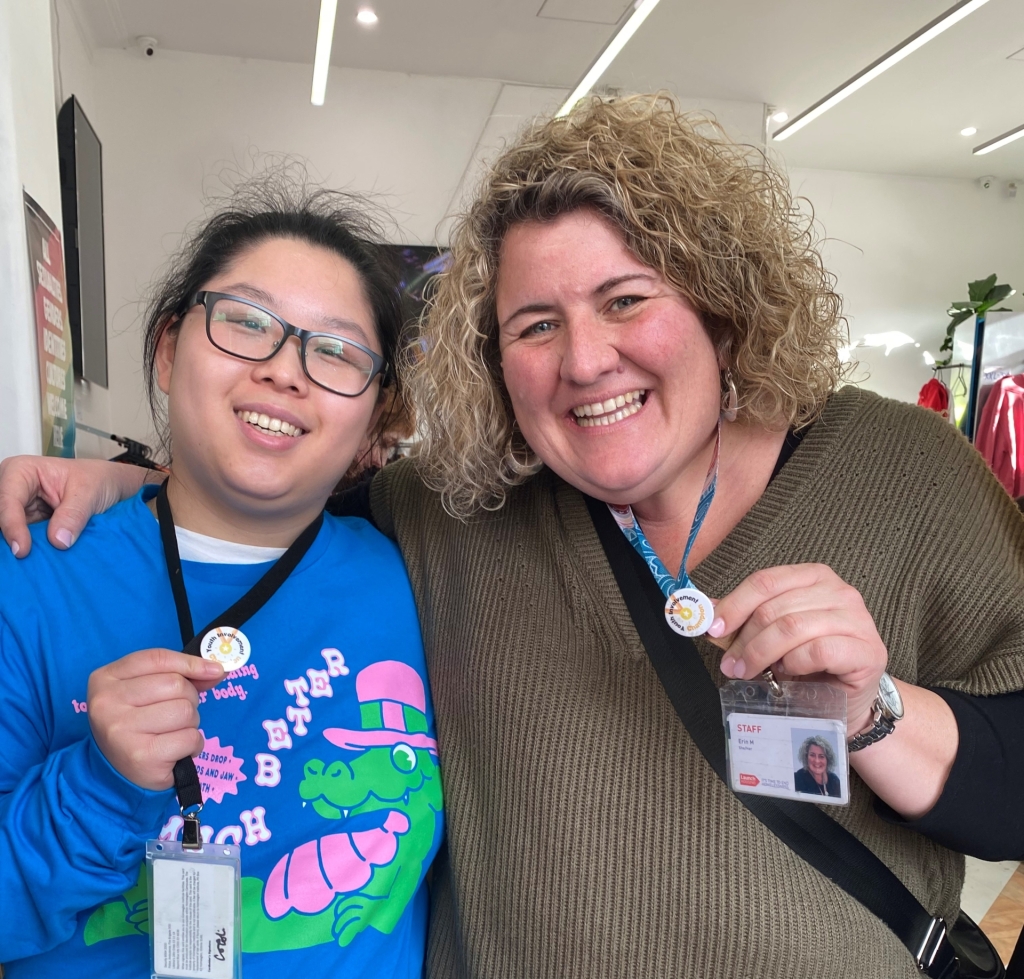 Two women (one a student and the other a staff member from the Youth Foyer in Glen Waverley, Victoria. They are smiling at the camera, showing off badges that have been made for them by a young person in Tasmania.