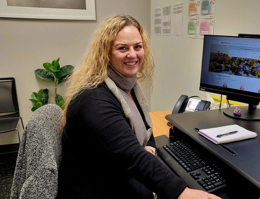 Hayley has a real estate website open on her computer. She is smiling at the camera. It is her job to help Tasmanians find a private rental property.