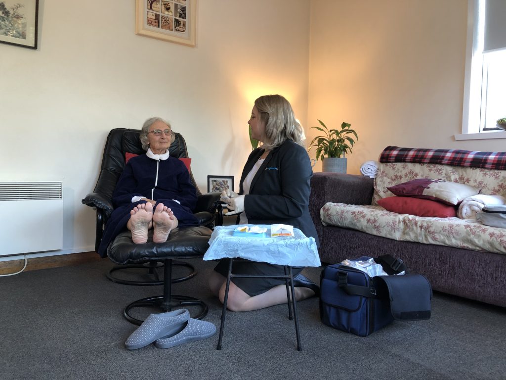 Two people in a lounge room. One person is wearing an Anglicare nurse uniform and is kneeling beside the other person who is sitting on a couch with their feet up on a stool They don't have any shoes on. It appears that the nurse is getting ready to change some dressings on a wound.