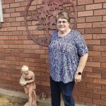 Jennifer Wallis at home standing beside her garden statue smiling.