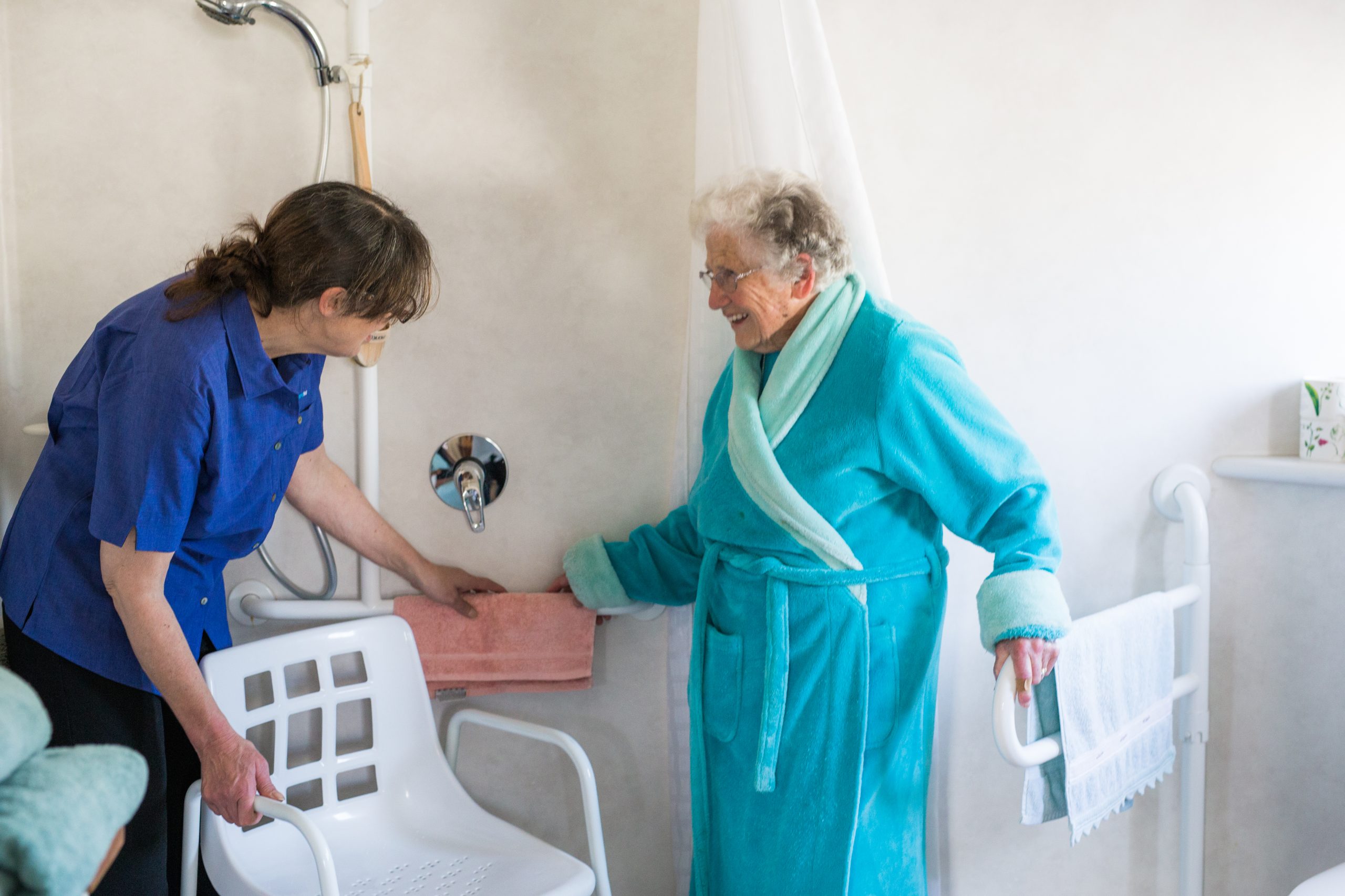 Two ladies are standing in a bathroom. One lady in a bathrobe getting ready to have a shower she is holding onto rails on the bathroom wall.. The lady wearing an Anglicare Home Care Support Worker Uniform is helping the lady in the robe turn on the shower and get into her shower chair. 