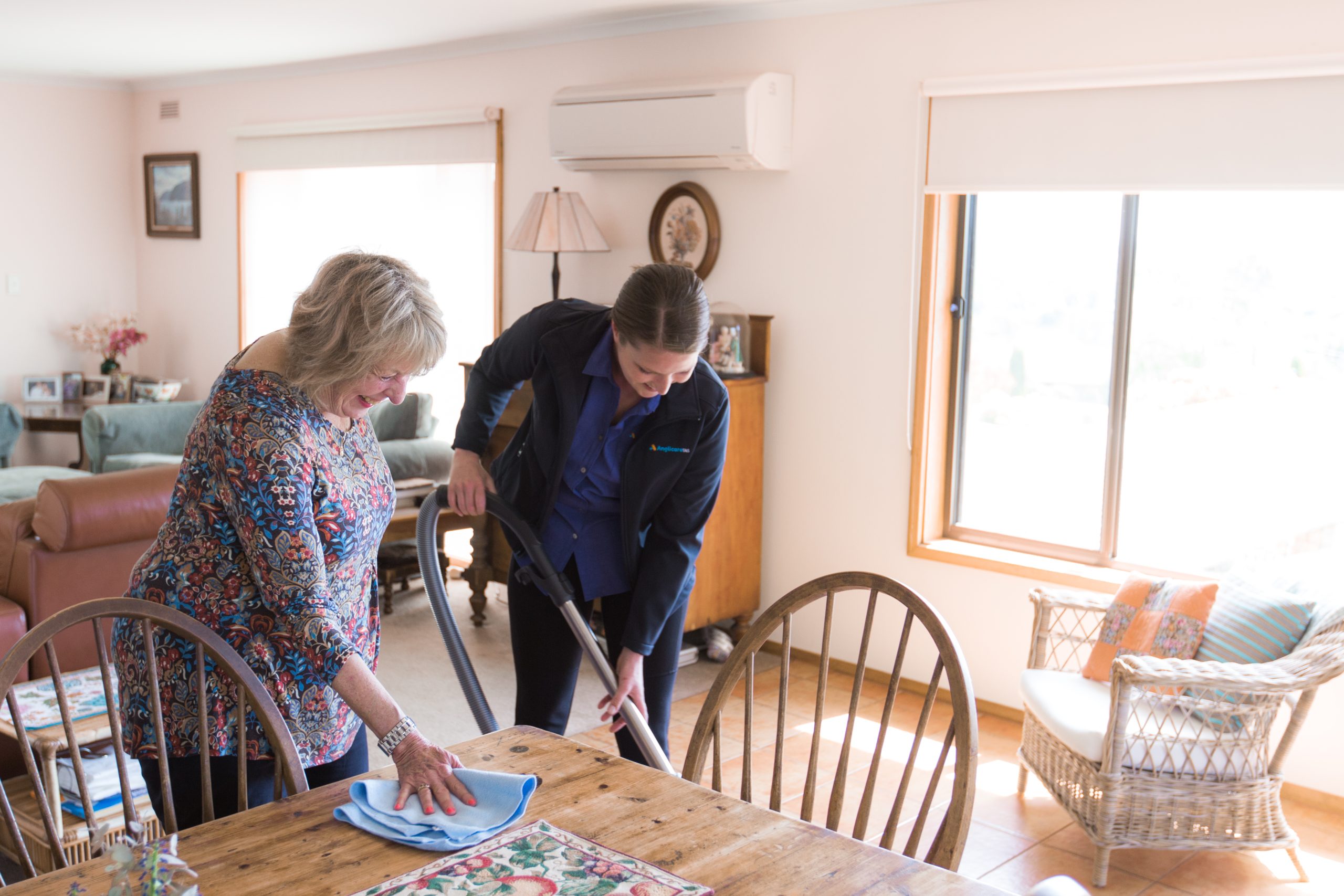 Two ladies doing housework together in a house. One is cleaning the tabletop and the other is vacumning. The person vacuuming is wearing an Anglicare Tasmania Home Care Support Worker uniform.