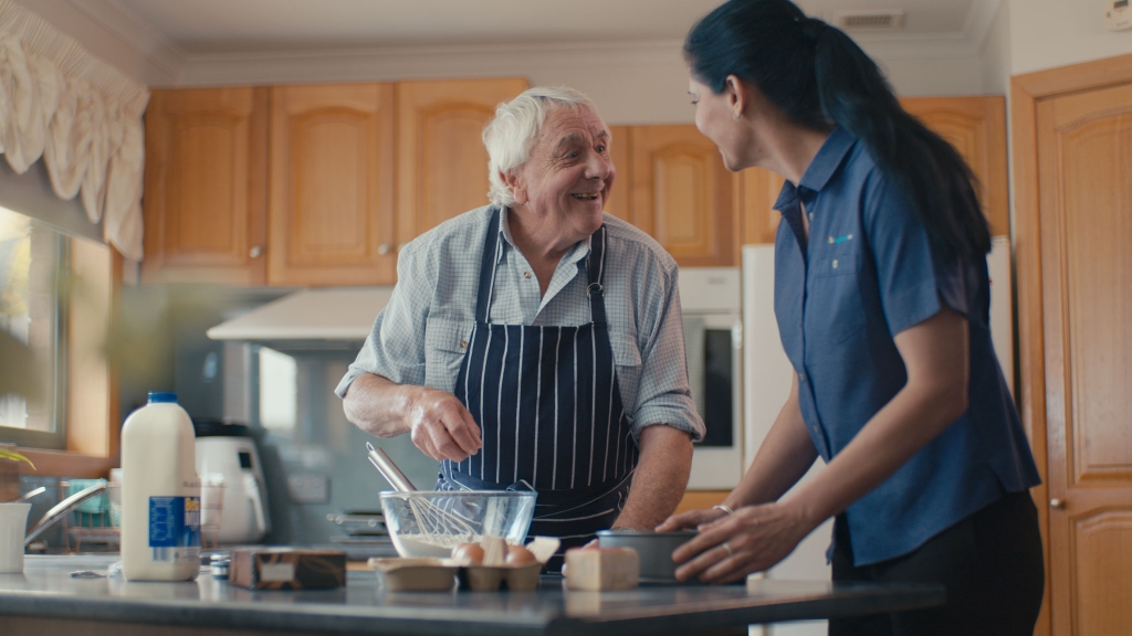 A man in the kitchen cooking and an Anglicare Support Worker speaking to him as he cooks.