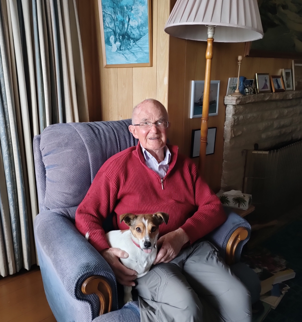 Older person, with glasses, sitting on a couch, wearing a red jumper and grey pants. They are holding a small white dog on their lap. They are smiling.